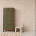 A green and wood dresser stands next to a small beige plastic chair on a checkered rug, set against a cream wall adorned with Elsies Verdant Garden Wallpaper.