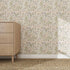 A light wood dresser with three drawers sits on a woven rug beside a wall covered in Forest Floor Wallpaper Repeat Pattern, featuring leaves and small flowers in soft, earthy tones.