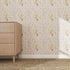 A wooden dresser with three drawers stands on a woven rug beside a wall featuring the Garden Symphony Wallpaper Repeat Pattern | Sample in soft pastel florals. The scene is brightly lit and minimalist.