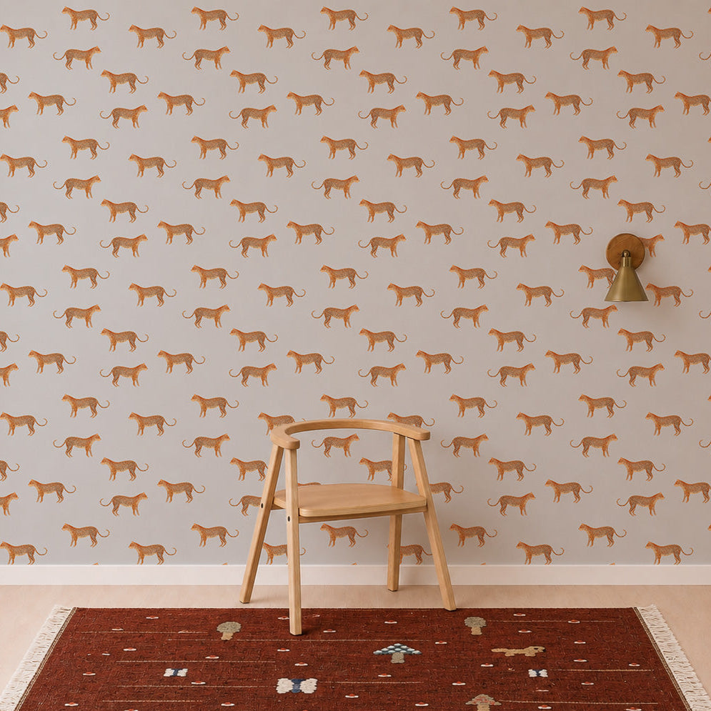 A wooden chair sits on a patterned rug in front of a wall adorned with Goldies Cheetah Wallpaper in Blue, featuring playful cheetahs and repeating orange leopard prints. A gold wall sconce is mounted to the right of the chair.