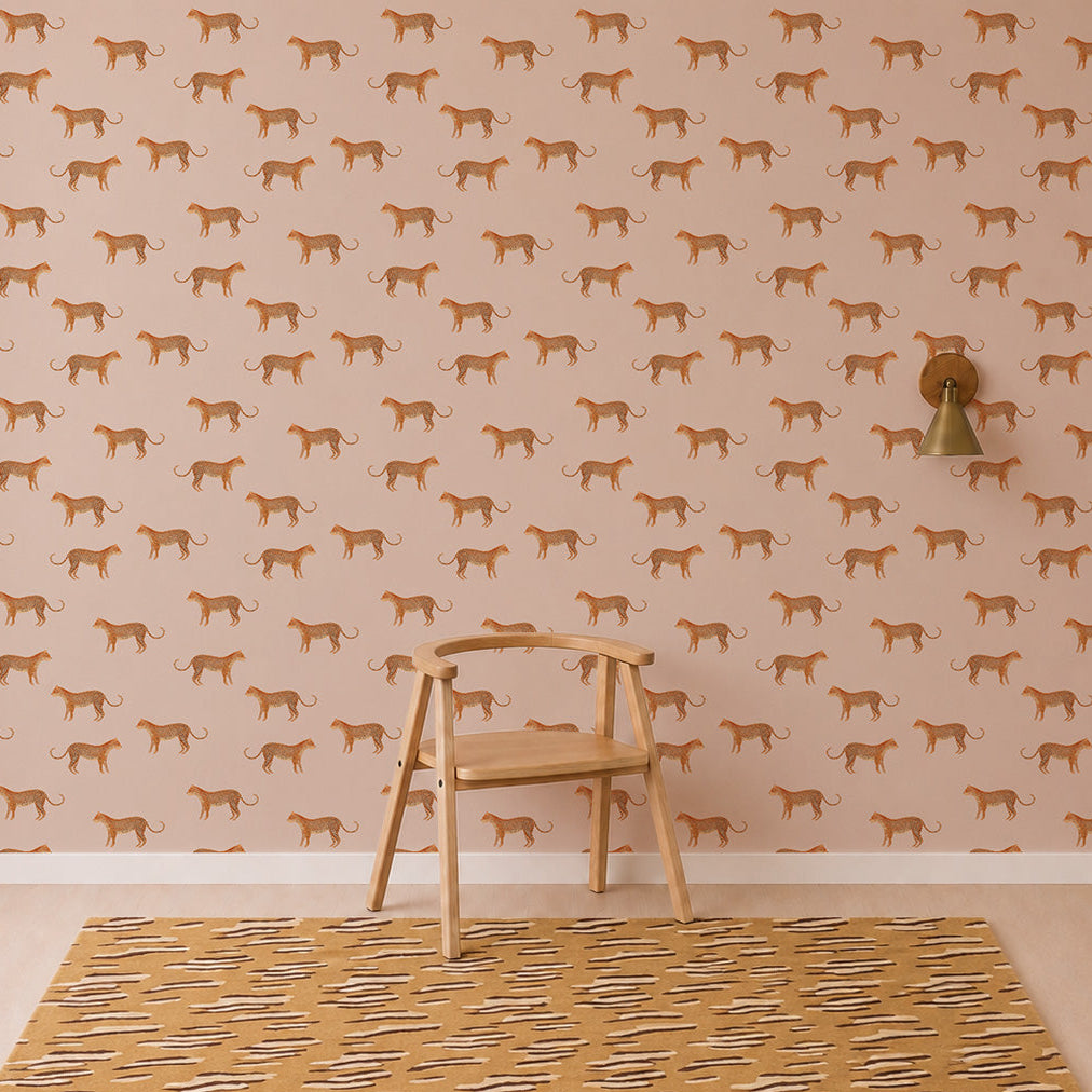 A wooden chair sits on a patterned rug in front of Goldies Cheetah Wallpaper in Pink, featuring a lively pattern of repeating orange cheetahs for a playful touch. A gold wall sconce is mounted to the right of the chair.
