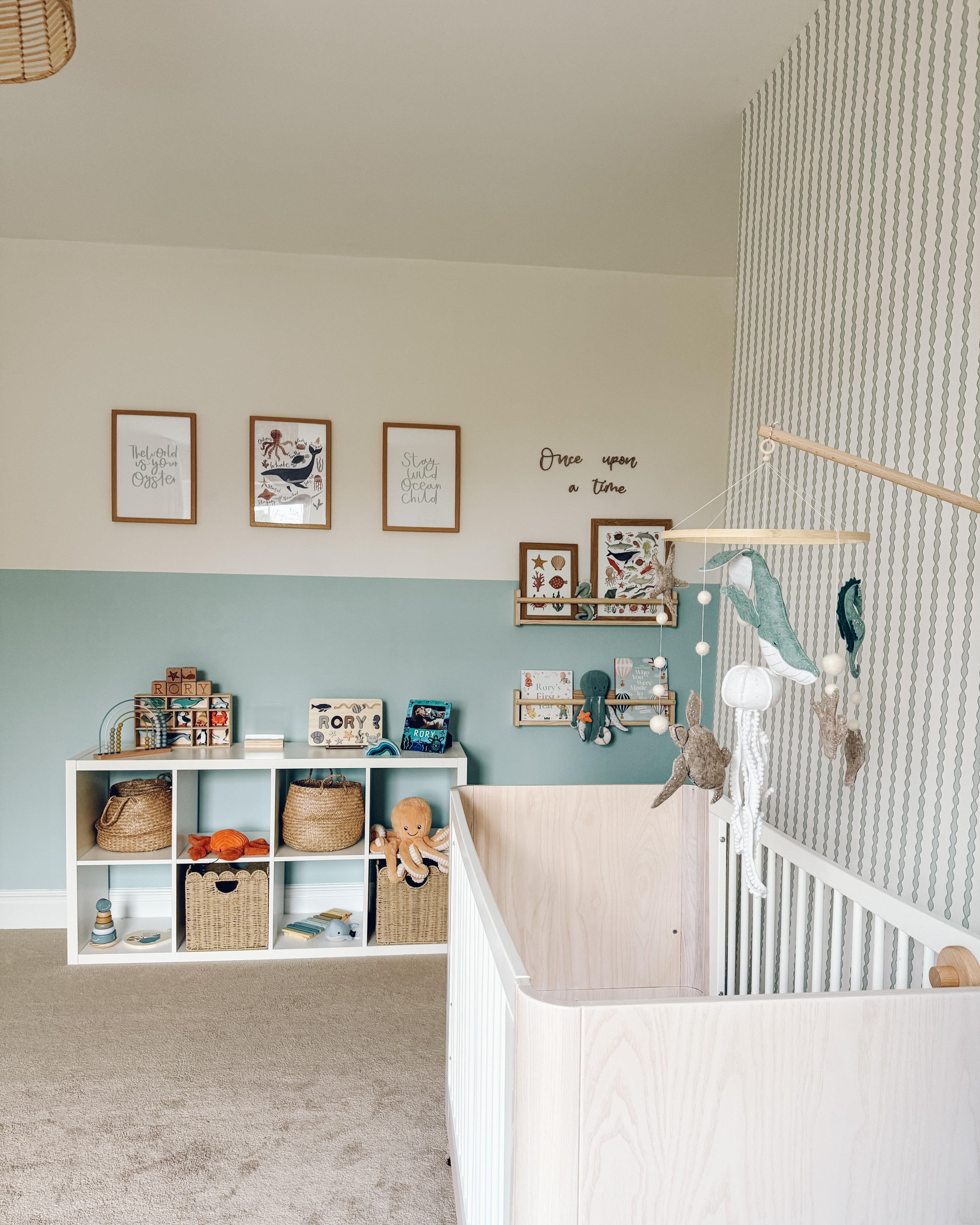 A cozy nursery with a light wooden crib, plush animals, and books features Little Abacus Stripe Blue and Green wallpaper on an accent wall, cubby storage with toys and baskets, framed wall art, and a soft beige carpet.