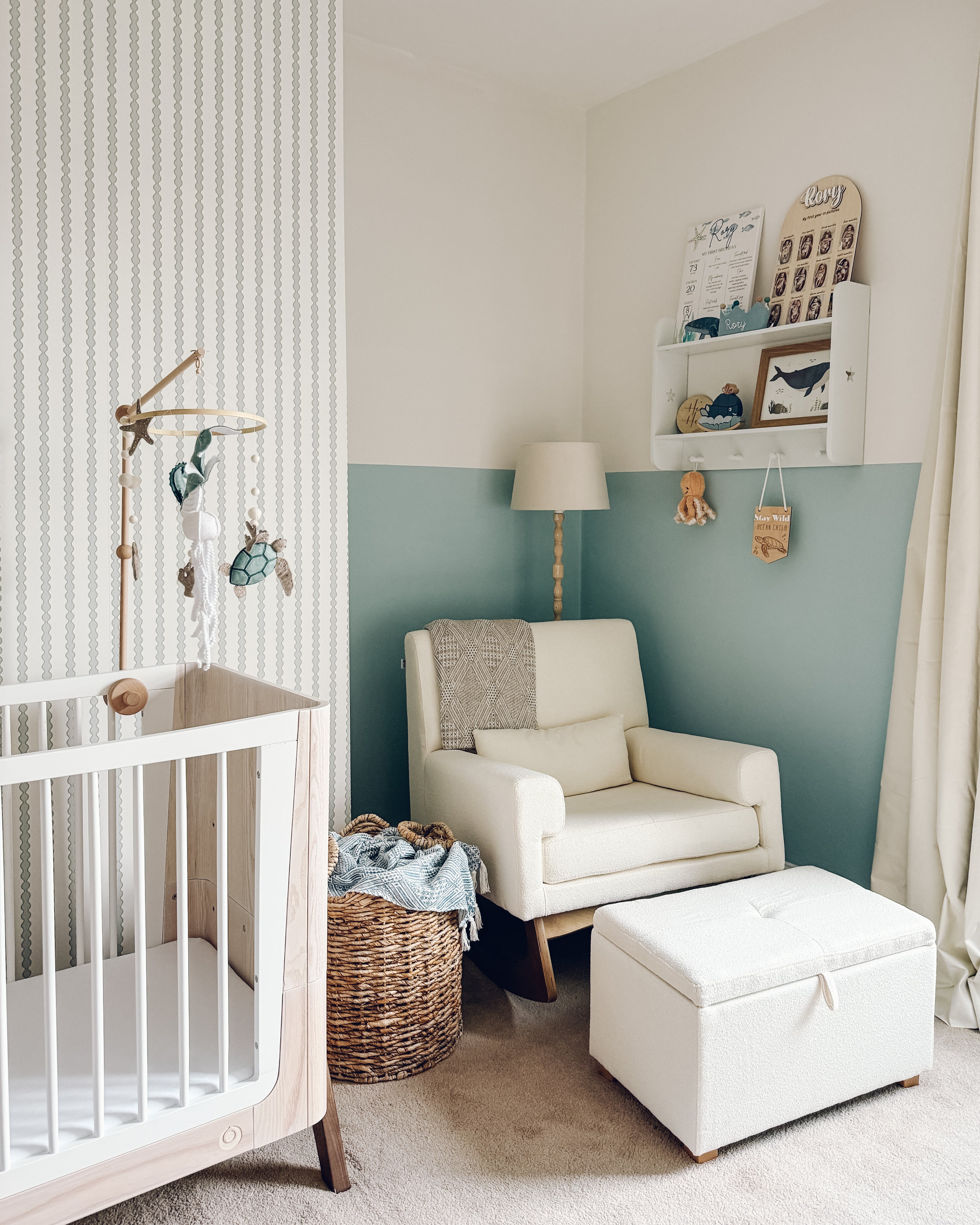 A cozy nursery features a white crib, a wicker basket, comfy armchair and ottoman, plus a floor lamp. Little Abacus Stripe Blue and Green wallpaper pairs perfectly with half-painted teal and white walls for added charm.