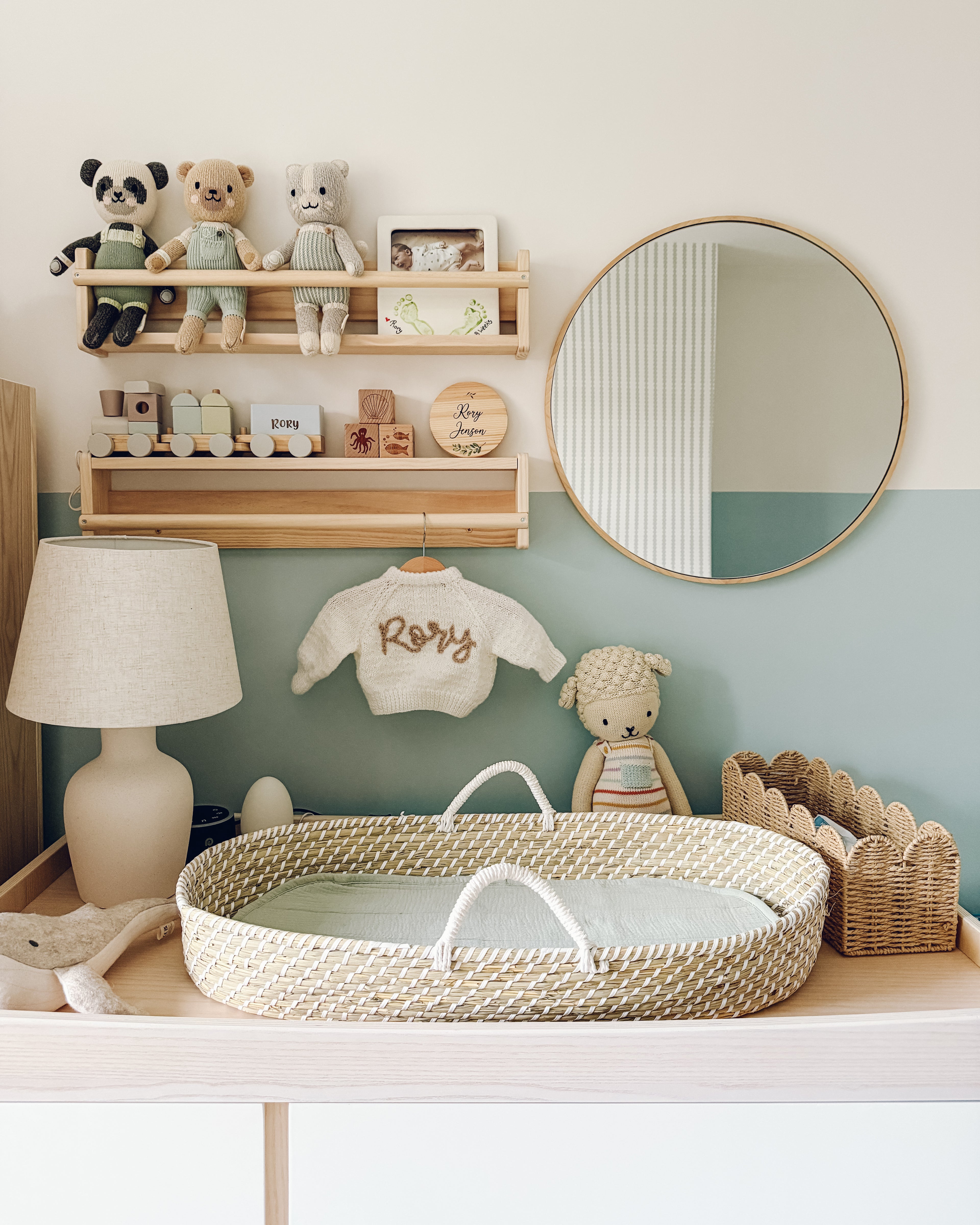A baby changing table with a woven basket, stuffed animals, baby items, a lamp, and a personalized "Rory" sweater sits below shelves and a round mirror on blue and green Little Abacus Stripe wallpaper.