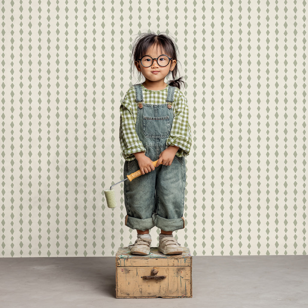 A young child in glasses, a green checked shirt, and denim overalls stands on a wooden box with a paint roller, set against vibrant Jolly Jester Wallpaper Repeat Sage Green.