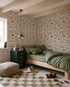 A cozy bedroom with green bedding on a wooden bed, a checkered rug, toys, books, and nursery walls decorated with Little Wanderers Wallpaper featuring colorful animals. Sunlight pours in through the window.