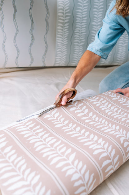 Wearing a blue shirt, a person uses large scissors and a ruler to cut Meadow Leaf Stripe Wallpaper, Clay—a botanical wallpaper with a white leaf pattern on beige—ideal for nurseries or hand-painted vine stripe designs.