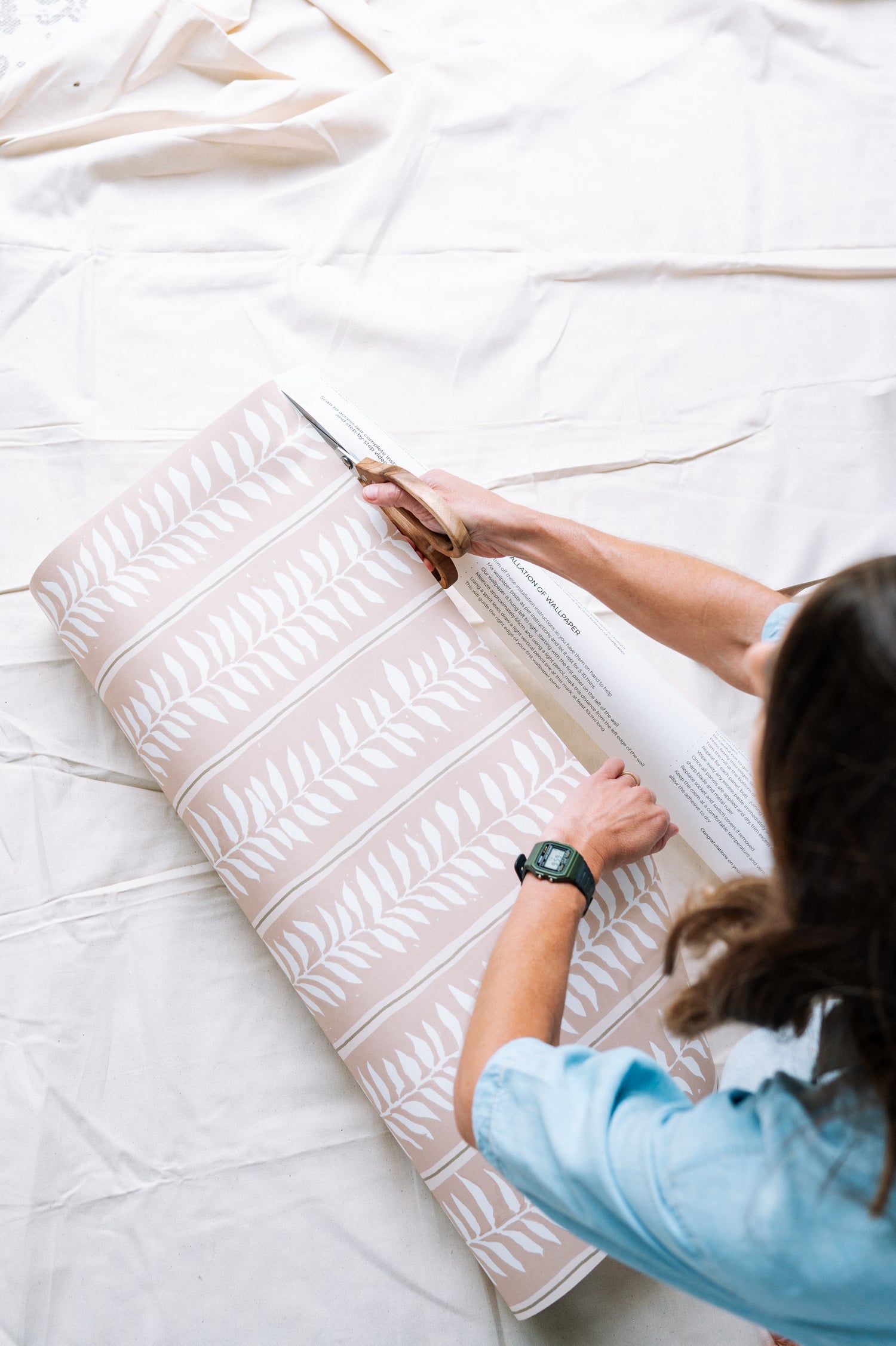 A person in a blue shirt and watch cuts a roll of Meadow Leaf Stripe Wallpaper, Clay, featuring pink botanical designs with white leaf patterns, on a white surface.