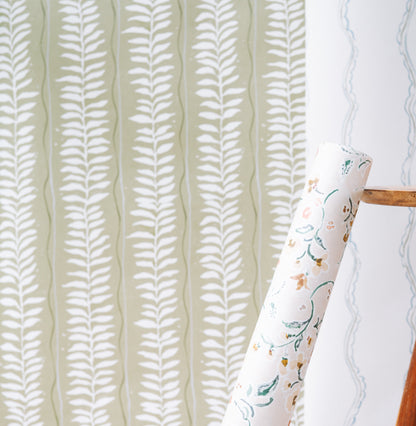 A roll of Meadow Leaf Stripe Wallpaper, Sage, sits on a wooden ladder before a wall covered in the same leafy pattern; to the right, another wall features a soft blue design.