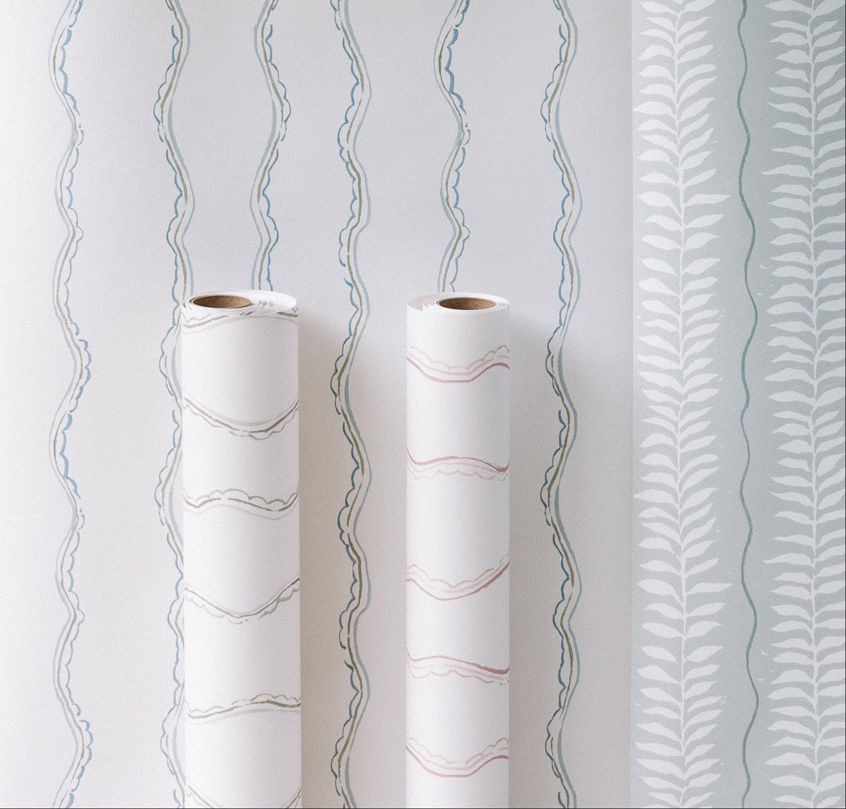 Two rolls of Ribbon Stripe Wallpaper, Blue, with wavy pastel patterns are displayed in front of a wall featuring blue wavy lines and white leafy designs on light blue—ideal for elegant children’s room decor.