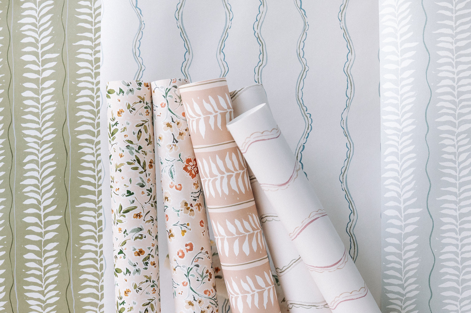 Four rolls of Meadow Leaf Stripe Wallpaper in Clay, featuring hand-painted botanical leaf patterns, stand upright against three background samples with matching leafy and wavy pastel designs.