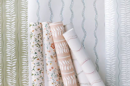Four rolls of Meadow Leaf Stripe Wallpaper in Clay, featuring hand-painted botanical leaf patterns, stand upright against three background samples with matching leafy and wavy pastel designs.