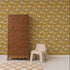 A brown wooden dresser and a small beige plastic chair sit on a light floor beside a tan-checkered rug, while the Mils Menagerie Wallpaper, Gold showcases deer and trees in vibrant yellow, green, and orange tones, bringing a playful touch of nature to the space.