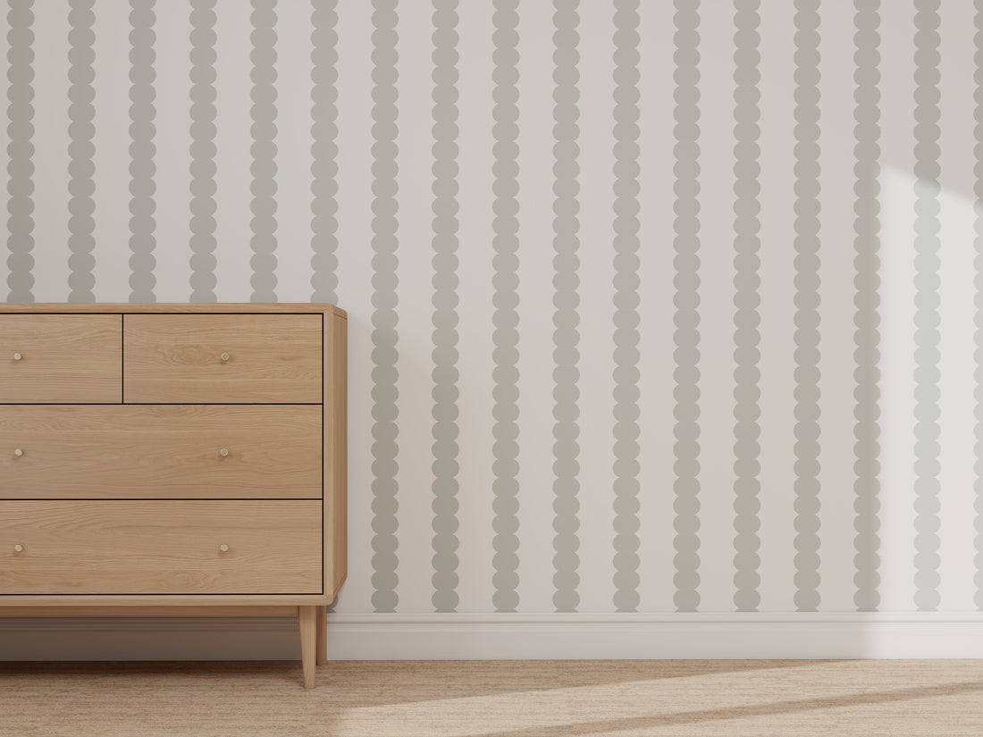 A light wooden dresser with four drawers stands against a wall featuring the Custom Scallop Stripe Wallpaper Blue sample. Sunlight illuminates the right side, and a light-colored rug covers the floor.