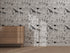 A light wooden dresser stands before the Going on Safari Wallpaper Repeat Pattern | Black and White, showcasing black-and-white giraffes, rhinos, elephants, leopards, and leaves as sunlight casts soft shadows on the illustrations.