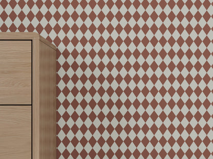 A light wood dresser stands against a wall covered in Diamond Dancer Wallpaper, Stone Rose, showcasing an artisanal pattern of red and white harlequin diamonds.