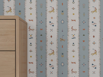 A light wooden cabinet stands beside a wall covered in The Little Carousel Stripe Wallpaper, Blue, which features blue and white stripes with whimsical animals, floral motifs, hearts, and charming decorative details.