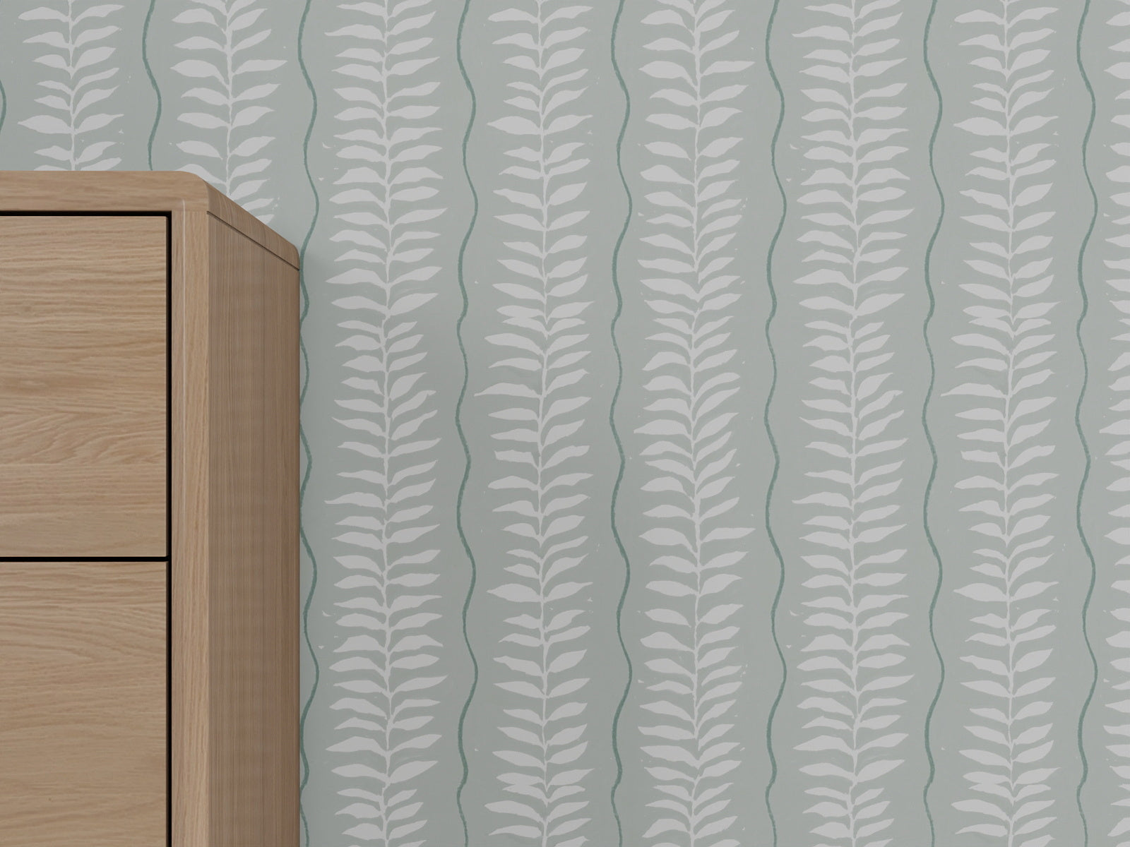 A light wooden cabinet stands against a wall decorated with Meadow Leaf Stripe Wallpaper in Sea Foam, featuring a greenish-gray background and vertical white botanical leaf pattern.