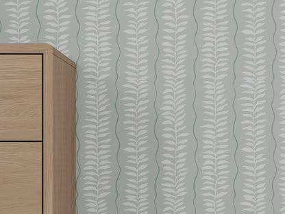 A light wooden cabinet stands against a wall decorated with Meadow Leaf Stripe Wallpaper in Sea Foam, featuring a greenish-gray background and vertical white botanical leaf pattern.