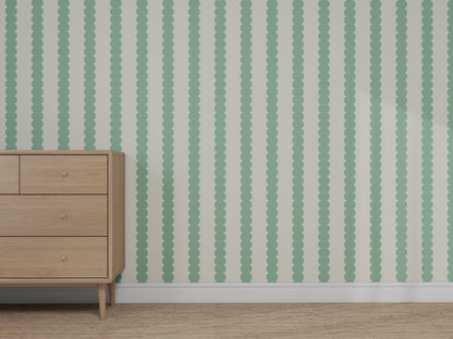 A light wood dresser stands in front of a wall covered with Custom Green Seafoam Scallop Stripe Wallpaper | Sample. The wallpaper features vertical mint green scalloped stripes on a light background, and the floor is carpeted in light tan.