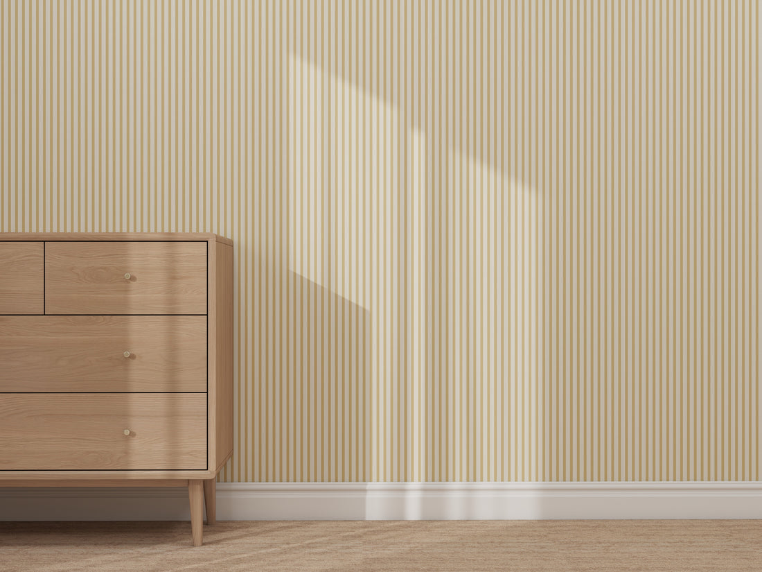 A wooden dresser with drawers stands on a beige carpet beside a wall decorated with Pico Stripe Wallpaper Yellow. Sunlight creates a rectangular shadow on the striped wallpaper, adding to the cozy and inviting atmosphere.