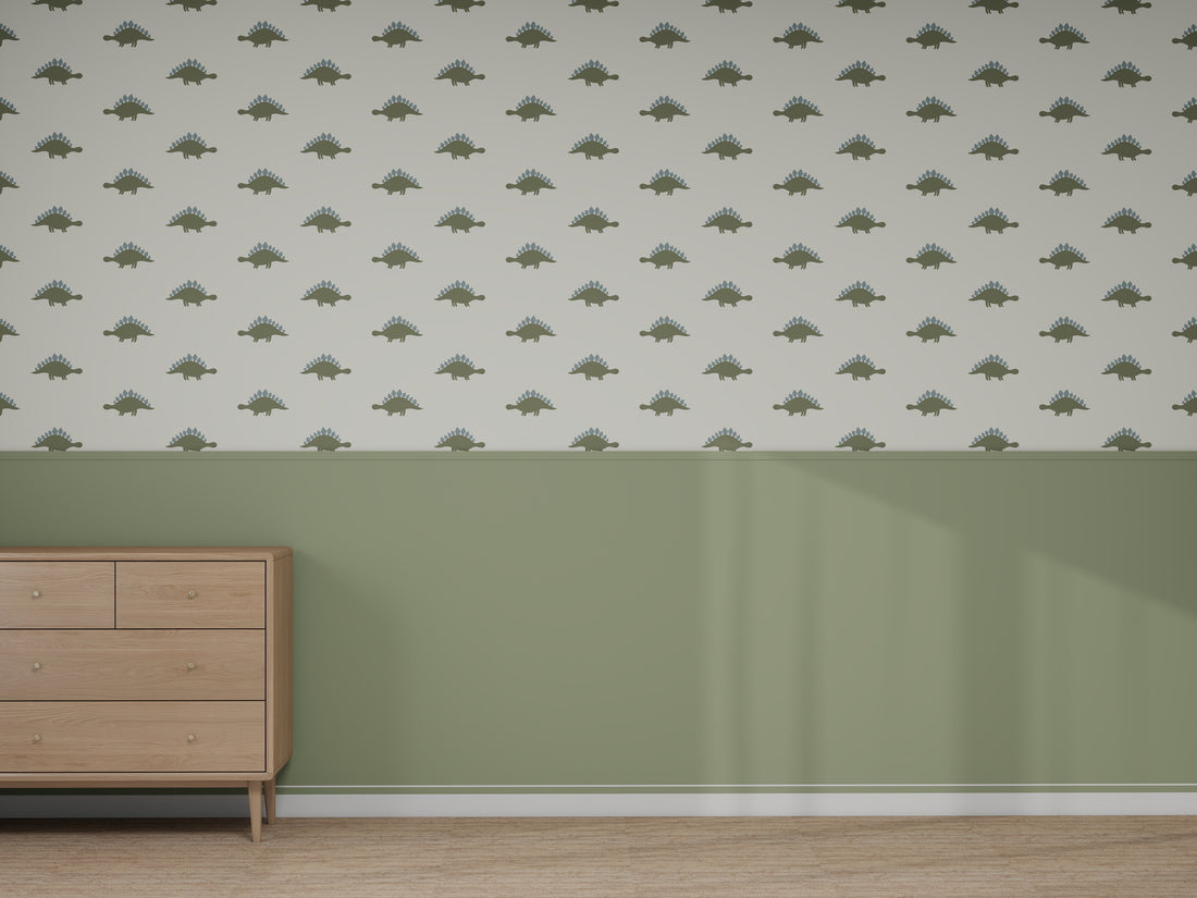 A room features light wood floors, sage green lower walls, and a "Custom Otis Dinosaur 2 | Sample" wallpaper with green dinosaur shapes above. A light wood dresser stands on the left as sunlight highlights the space’s colors and quality.