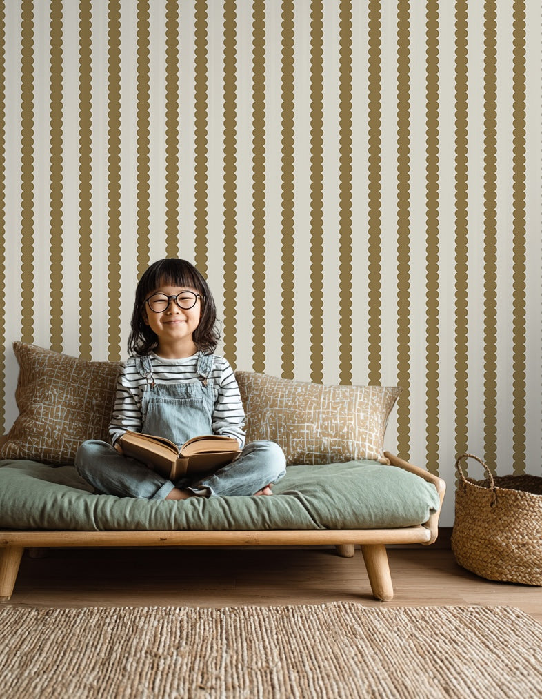 A smiling young girl with glasses and overalls sits cross-legged on a cushioned wooden bench, holding an open book. Decorative pillows and a basket are nearby, while Scallop Stripe Wallpaper Moss brings charm to the cozy kids&