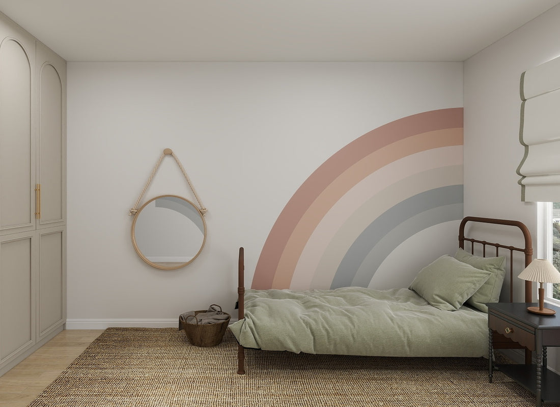 A cozy childrens space featuring a single bed with sage green bedding, a round mirror, woven rug, basket, small dark nightstand with lamp, and the Corner Rainbow Wallpaper Mural as a pastel feature wall.