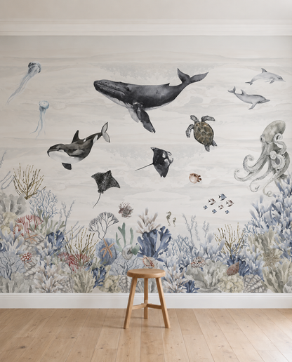 A wooden stool sits before the Ocean Magic Mural, Watercolour, featuring a whale, turtle, and octopus above vibrant coral reefs. This underwater wallpaper brings a calming and artistic touch to any kids room.