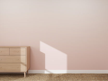 A minimalist room featuring the Ombre Wallpaper Mural | Pink, a light wooden dresser on the left, sunlight casting window-shaped shadows, and a woven beige rug on the floor.