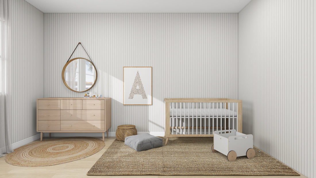 A minimalist nursery with Blush Rose accents showcases a wooden crib, dresser with round mirror, woven rug, and toy cart on textured carpet. munks and mes Pinstripe Wallpaper Soft Beige adds charm with framed letter A art on the wall. Soft cushions are nearby, and the room is softly lit.