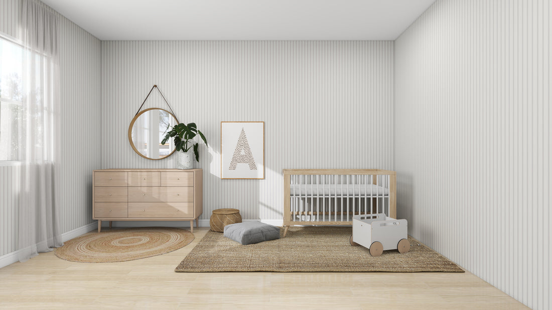 In a minimalist nursery with light walls and wood floors, munks and mes Pinstripe Wallpaper in Soft Beige stands out. The space includes a crib, dresser with round mirror, geometric wall art, blush rose rug, toy box on wheels, and warm light filtering through the window.