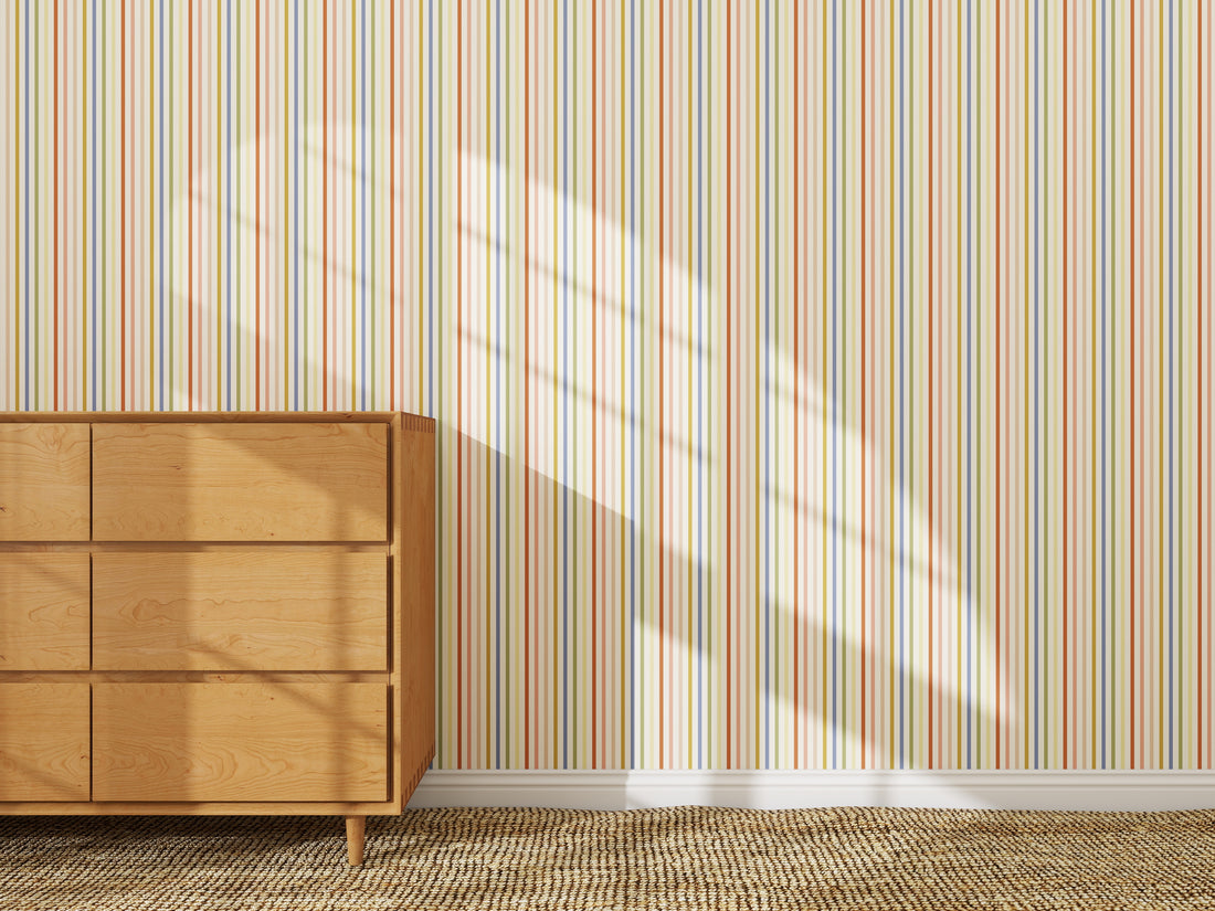 A wooden dresser stands against a wall decorated with Pico Stripe Wallpaper Multi. Sunlight casts window-shaped shadows on the vibrant striped wallpaper, while a textured woven rug covers the floor.