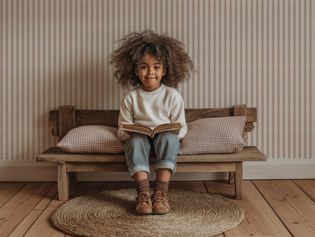 A young child with curly hair smiles while reading on a wooden bench with cushions. Behind them, the nursery has Little Abacus Stripe Rose wallpaper, and a round woven rug lies on the wooden floor.