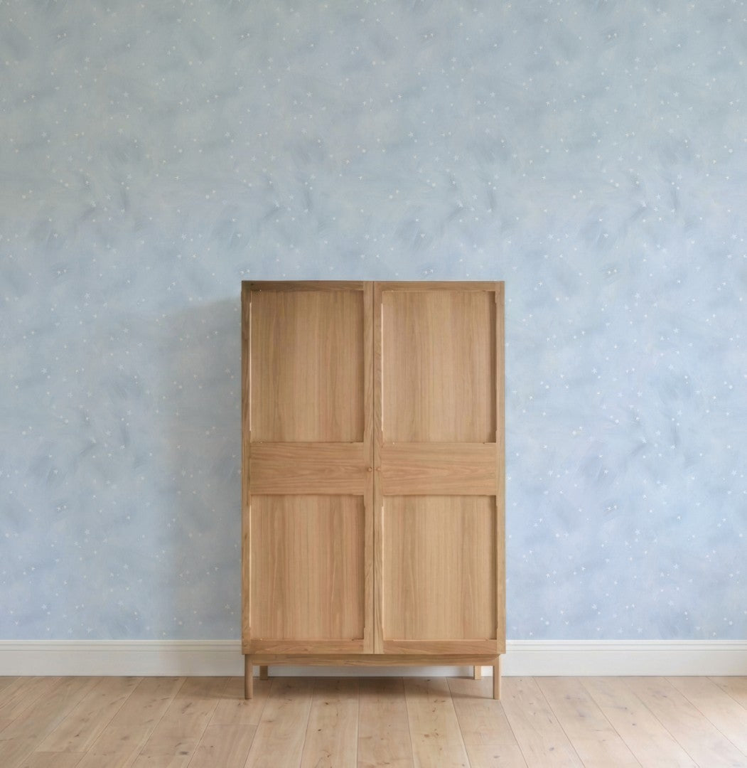 A simple wooden cabinet with four panels stands against a light wood floor and a wall decorated with Painted Cloud Stars Wallpaper, Blue, featuring subtle white speckles for a celestial effect.