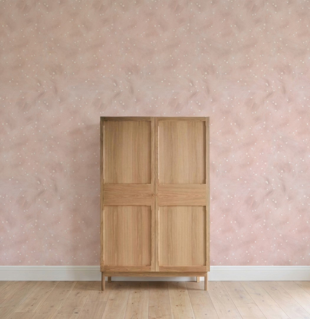 A wooden wardrobe with four panels stands against a pink wall adorned with Painted Cloud Stars Wallpaper, Rose. The light wood floor and minimal decor create an uncluttered, serene nursery setting.