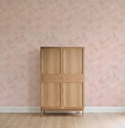 A wooden wardrobe with four panels stands against a pink wall adorned with Painted Cloud Stars Wallpaper, Rose. The light wood floor and minimal decor create an uncluttered, serene nursery setting.