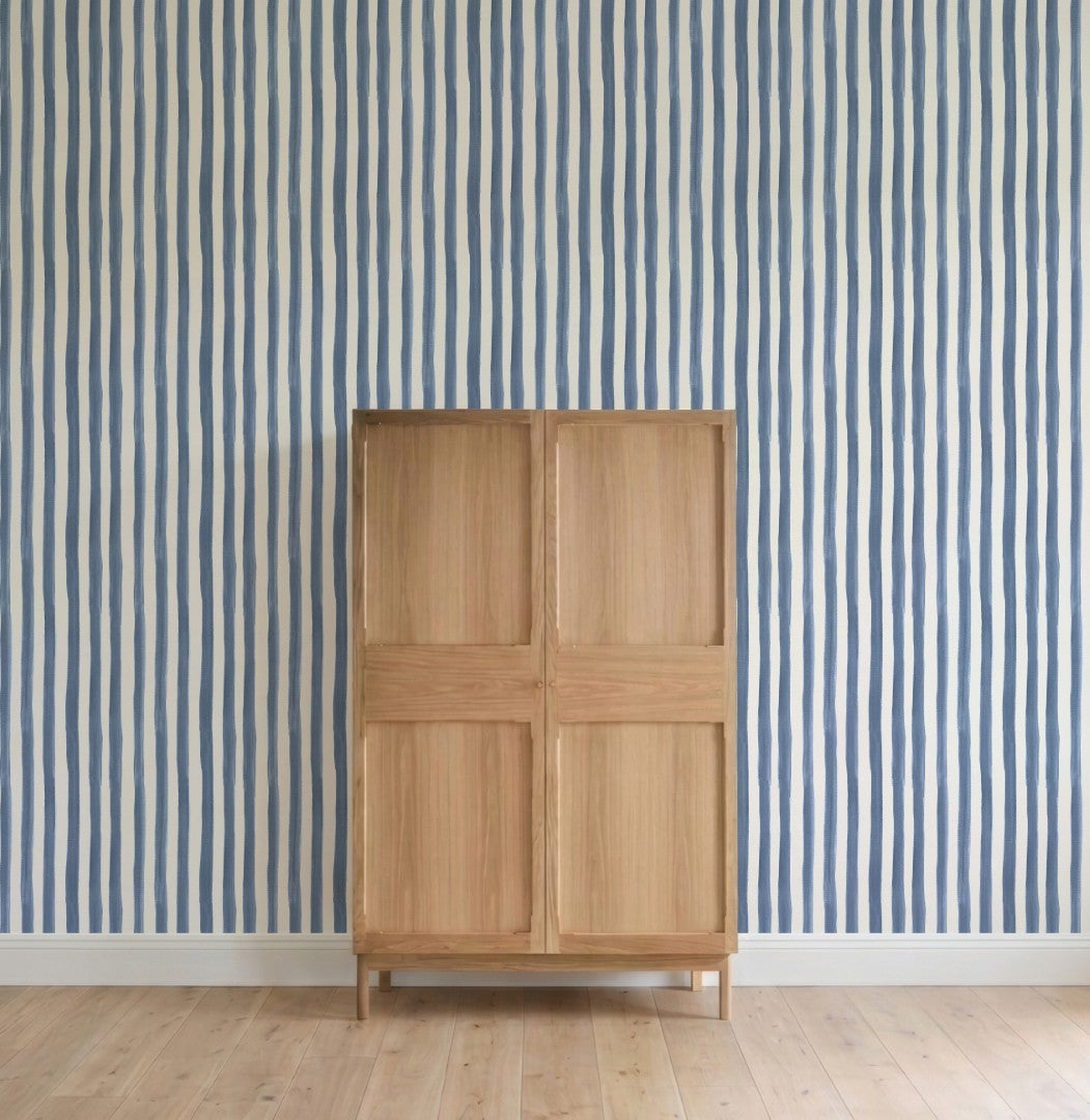 A light wooden cabinet stands on a wooden floor before a wall adorned with Painted Stripe Wallpaper, Blue. The scene is minimalist, displaying only the striped wallpaper and the cabinet without additional décor.