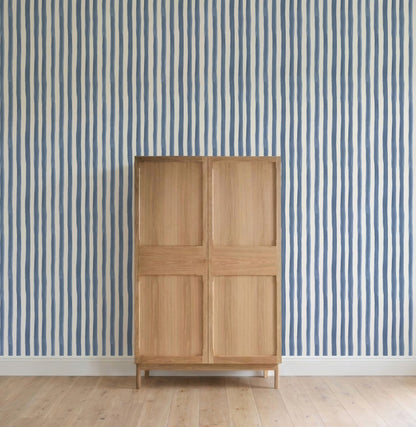 A light wooden cabinet stands on a wooden floor before a wall adorned with Painted Stripe Wallpaper, Blue. The scene is minimalist, displaying only the striped wallpaper and the cabinet without additional décor.