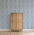A light wooden cabinet stands on a wooden floor before a wall adorned with Painted Stripe Wallpaper, Blue. The scene is minimalist, displaying only the striped wallpaper and the cabinet without additional décor.