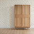 A light wooden wardrobe with two doors stands on a light wood floor, backed by a wall featuring Soaring Hearts Wallpaper Sample—its colorful floral pattern highlights excellent quality in this minimalistic, tidy room.
