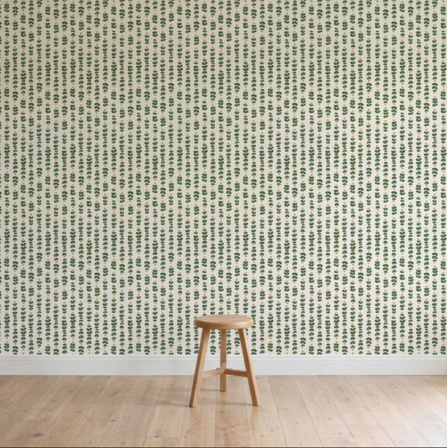 A simple wooden stool sits on a light wood floor in front of a wall adorned with Floral Trails Wallpaper, displaying vertical rows of small green leafy plants on a crisp white background.