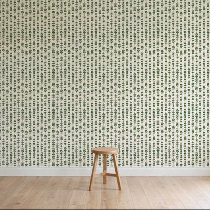 A simple wooden stool sits on a light wood floor in front of a wall adorned with Floral Trails Wallpaper, displaying vertical rows of small green leafy plants on a crisp white background.