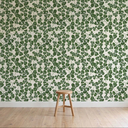 A wooden stool stands on a light wood floor in front of a wall covered with How Lucky We Are Clovers Wallpaper, featuring green leafy hand-painted designs.