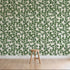 A wooden stool stands on a light wood floor in front of a wall covered with How Lucky We Are Clovers Wallpaper, featuring green leafy hand-painted designs.