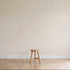 A simple wooden stool stands on a light wood floor against a plain wall with Jacquard Stars Wallpaper in gold, adding subtle elegance to the setting.