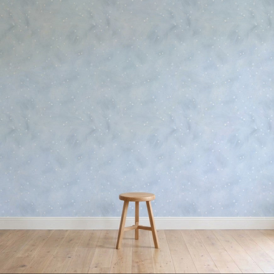 A single wooden stool sits on a light floor against Painted Cloud Stars Wallpaper, Blue—a pale celestial design with soft stars. The minimalistic room feels serene and empty.