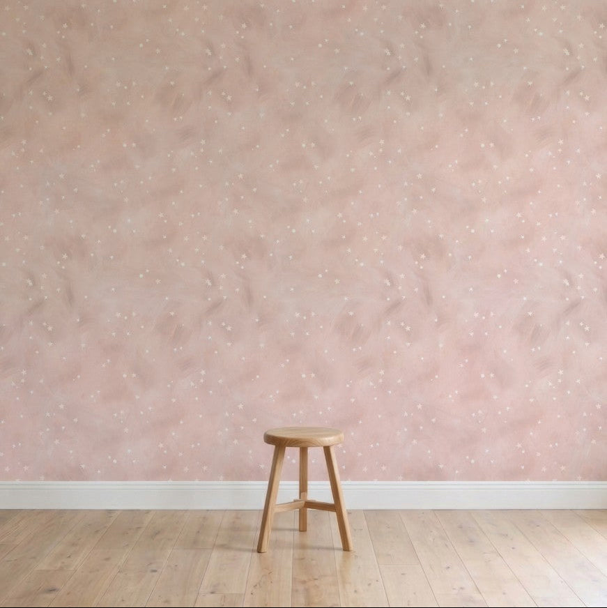 A single wooden stool sits on a light wooden floor against a pale pink wall decorated with Painted Cloud Stars Wallpaper, Rose, featuring subtle celestial patterns and white speckles in a softly lit, minimalistic scene.