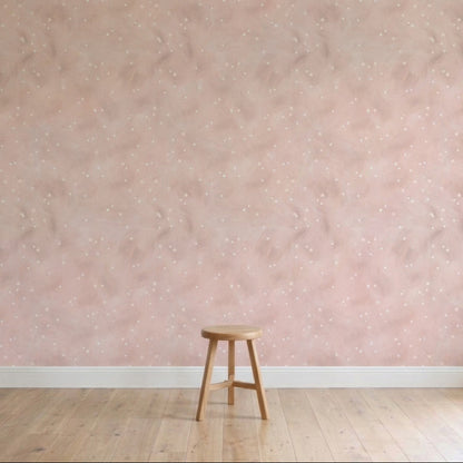 A single wooden stool sits on a light wooden floor against a pale pink wall decorated with Painted Cloud Stars Wallpaper, Rose, featuring subtle celestial patterns and white speckles in a softly lit, minimalistic scene.