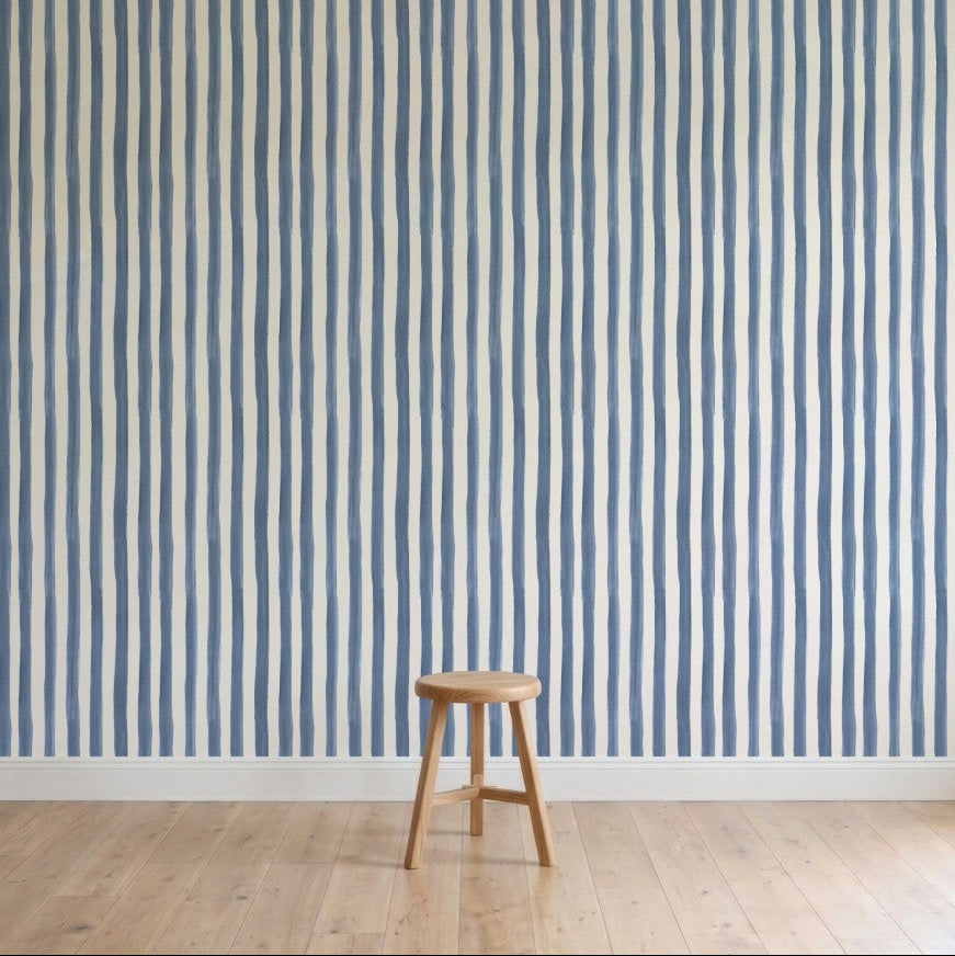 A simple wooden stool sits on a light wooden floor in front of a wall covered with Painted Stripe Wallpaper, Blue, featuring hand-painted vertical blue and white irregular stripes.